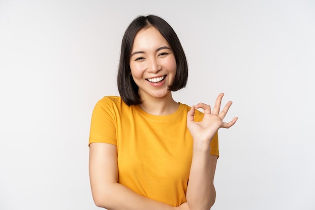 Schöne junge Frau, die ein okayzeichen zeigt, das erfreut lächelt und etw empfiehlt, genehmigen wie ein Produkt, das in einem gelben T-Shirt auf weißem Hintergrund steht