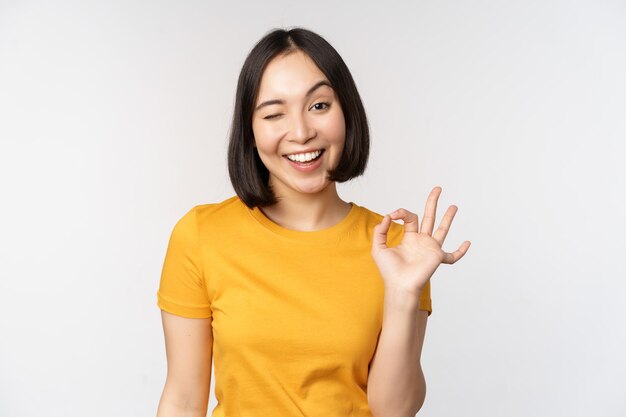 Schöne junge Frau, die ein okayzeichen zeigt, das erfreut lächelt und etw empfiehlt, genehmigen wie ein Produkt, das in einem gelben T-Shirt auf weißem Hintergrund steht