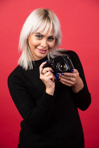 Schöne junge Frau, die eine Kamera hält, während gegen roten Hintergrund steht. Hochwertiges Foto