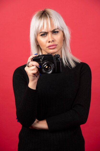 Schöne junge Frau, die eine Kamera hält, während gegen roten Hintergrund steht. Hochwertiges Foto