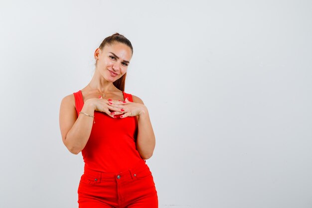 Schöne junge Frau, die gefaltete Finger auf Brust in rotem Trägershirt, Hosen hält und fröhlich schaut, Vorderansicht.