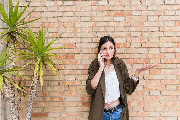 Schöne junge Frau, die gegen die Backsteinmauer spricht auf dem Mobiltelefonzucken steht