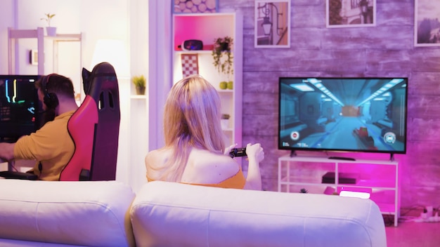 Schöne junge Frau, die glücklich ist, bei Online-Videospielen zu gewinnen, die auf dem Sofa mit drahtlosem Controller sitzen. Freund sitzt auf Gaming-Stuhl.