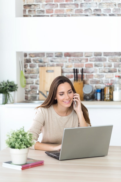 Schöne junge Frau, die Handy und Laptop in der Küche verwendet.