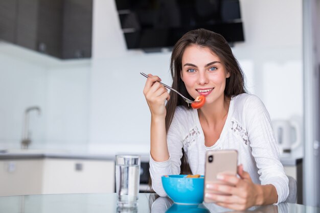 Schöne junge Frau, die Handy verwendet, während Salat in der Küche macht. Gesundes Essen. Gemüsesalat. Diät. Gesunder Lebensstil. Zu Hause kochen.