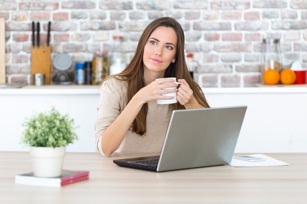 Schöne junge Frau, die ihren Laptop in der Küche verwendet.