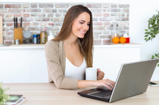 Schöne junge Frau, die ihren Laptop in der Küche verwendet.