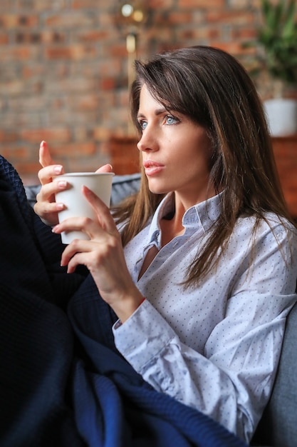 Schöne junge Frau, die im Sofa mit einem heißen Getränk entspannt