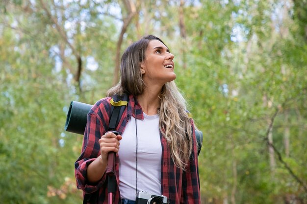 Schöne junge Frau, die in den Bergen mit Rucksack wandert. Aufgeregter weiblicher Reisender, der sich umschaut und lächelt. Grün auf Hintergrund. Backpacking Tourismus, Abenteuer und Sommerurlaub Konzept