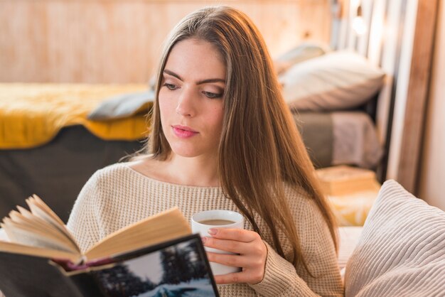 Schöne junge Frau, die Kaffeetasse-Lesebuch hält