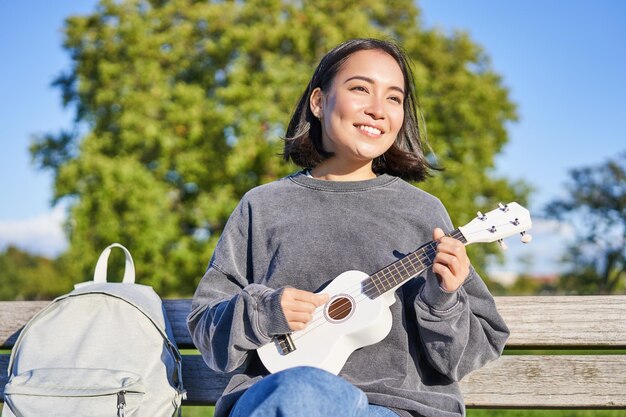 Schöne junge Frau, die mit Rucksack auf der Bank im sonnigen Park sitzt, spielt Ukulele-Gitarre und singt mit