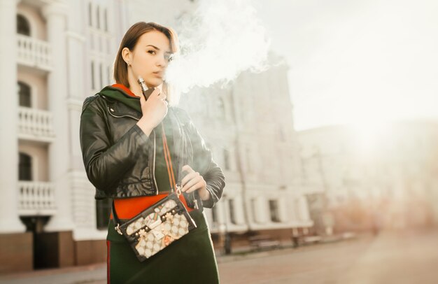 Schöne junge Frau, die Rauch inhaliert. junges Mädchen vaping vor dem Hintergrund der Stadt.