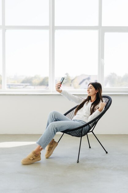 Schöne junge Frau, die sich mit ihrem Telefon selbst macht und lächelt, während sie zu Hause auf einem großen Stuhl sitzt