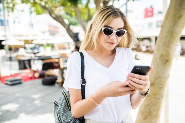 Schöne junge Frau, die Smartphone im Freien in der sonnigen Sommerstraße verwendet