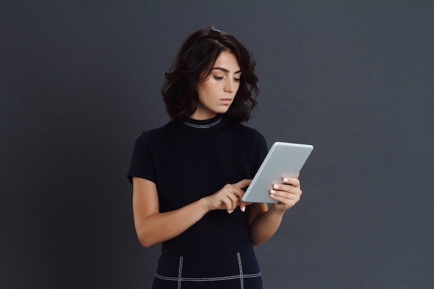 Schöne junge Frau, die über graue Wand aufwirft und Tablette in Händen hält