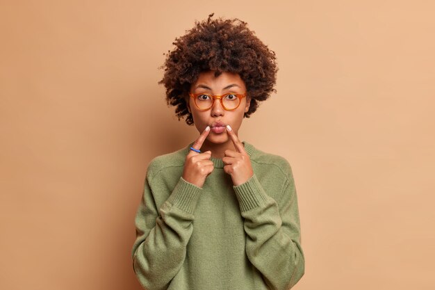 Schöne junge Frau hält Zeigefinger in der Nähe von gefalteten Lippen macht Gesicht vorne will küssen jemand trägt Brille und Pullover isoliert über braune Wand