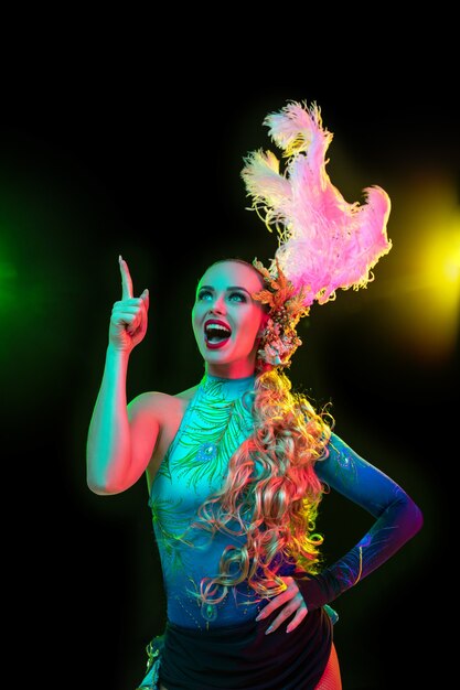Schöne junge Frau im Karneval, stilvolles Maskeradenkostüm mit Federn auf schwarzer Wand im Neonlicht