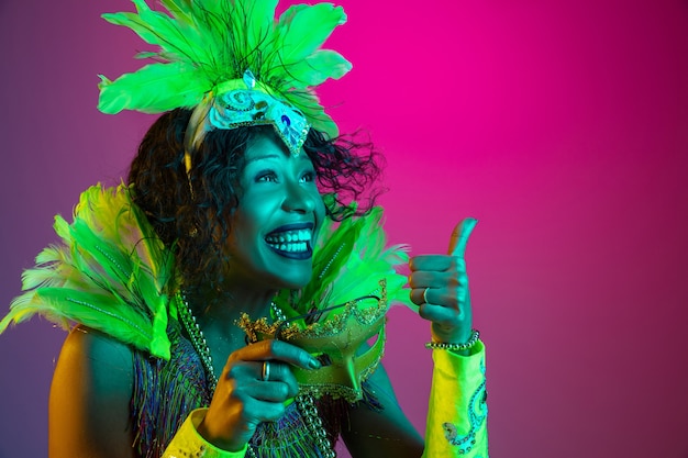 Schöne junge Frau im Karneval, stilvolles Maskeradenkostüm mit Federn, die auf Gradientenwand in Neon tanzen