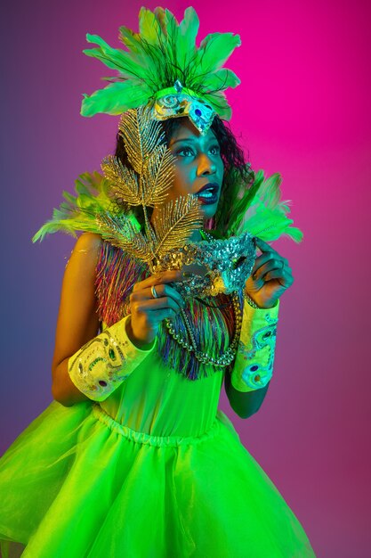 Schöne junge Frau im Karneval, stilvolles Maskeradenkostüm mit Federn, die auf Gradientenwand in Neon tanzen