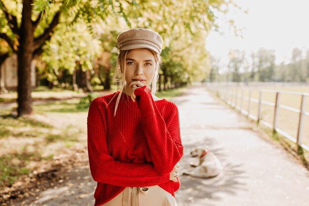 Schöne junge Frau im roten Pullover und im schönen trendigen Hut, der im Herbstpark nachdenklich aussieht. Attraktive Blondine in stilvoller Kleidung, die im Freien aufwirft.
