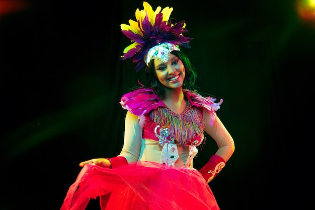 Schöne junge Frau in Karnevalsmaske und stilvollem Maskenkostüm mit Federn in bunten Lichtern und Leuchten an schwarzer Wand