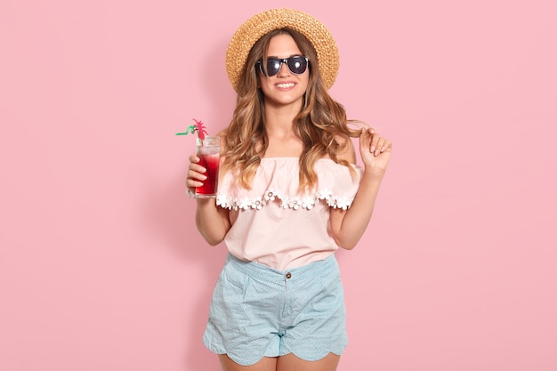 Schöne junge Frau in Sommerbluse, blaue kurze, schwarze Sonnenbrille und Strohhut, die Glas halten