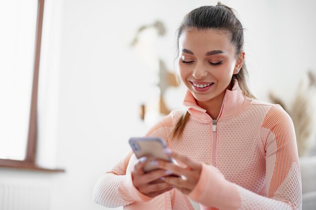 Schöne junge Frau in Sportkleidung, die drinnen sitzt und vor dem Training auf das Smartphone schaut