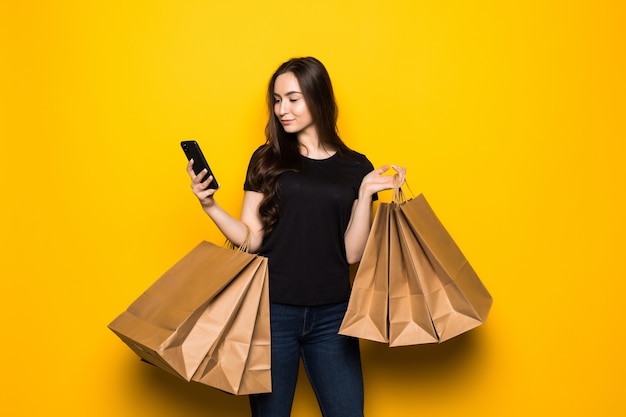 Kostenloses Foto schöne junge frau mit einkaufstaschen unter verwendung ihres smartphones auf gelber wand. shopaholic shopping mode.