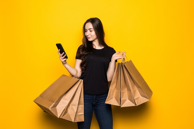 Schöne junge Frau mit Einkaufstaschen unter Verwendung ihres Smartphones auf gelber Wand. Shopaholic Shopping Mode.