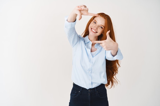 Schöne junge Frau mit langem Ingwerhaar und Sommersprossen, die durch Handrahmen schaut, den Moment einfängt oder nach dem perfekten Winkel sucht, über weißer Wand stehend