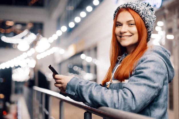 Schöne junge Frau mit Smartphone auf der nächtlichen Stadtstraße voller Licht. Porträt der herrlichen lächelnden Frau, die Handy verwendet.