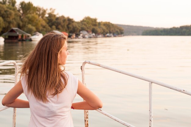 Schöne junge Frau schaut auf Sonnenuntergang über dem Fluss Attraktive Frau genießt Sommer und warmes Sonnenlicht. Sie bewundern schöne Aussicht und genießen Sonnenuntergang Sonnenlicht. Der Wind blies ihr Haar.