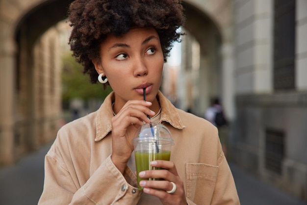 Schöne junge Frau trinkt Bio-Grün-Smoothie verwendet Plastikstroh, konzentriert in die Ferne, gekleidet in beige Jacken-Posen vor verschwommenem Hintergrund im Freien. Gesundes Getränk und Leute