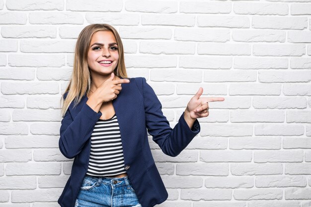 Schöne junge Frau über weiße Backsteinmauer, die mit glücklichem Gesicht lächelt und zur Seite zeigt