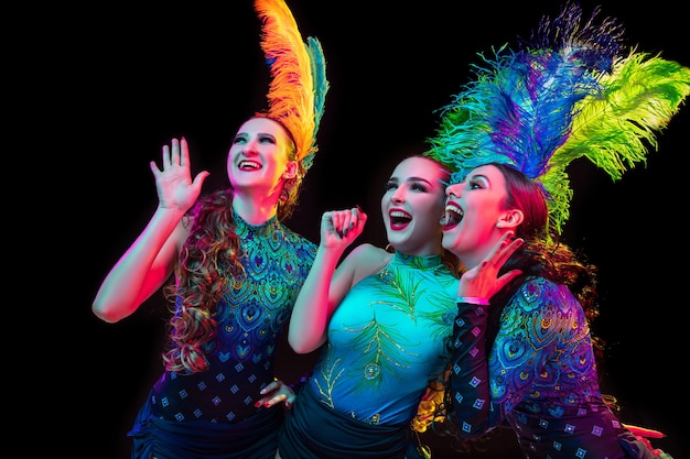 Schöne junge Frauen im Karneval, stilvolles Maskeradenkostüm mit Federn auf schwarzem Hintergrund im Neonlicht.