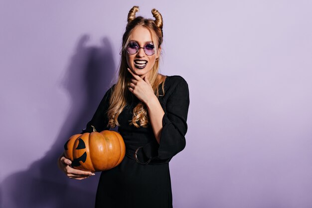 Schöne junge Hexe, die an lila Wand aufwirft. Glückseliges Vampirmädchen, das Halloween-Kürbis hält.