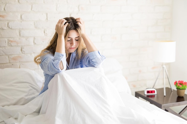 Schöne junge hispanische Frau, die ihr Haar hält und sich gestresst und besorgt fühlt, während sie zu Hause in einem Bett sitzt