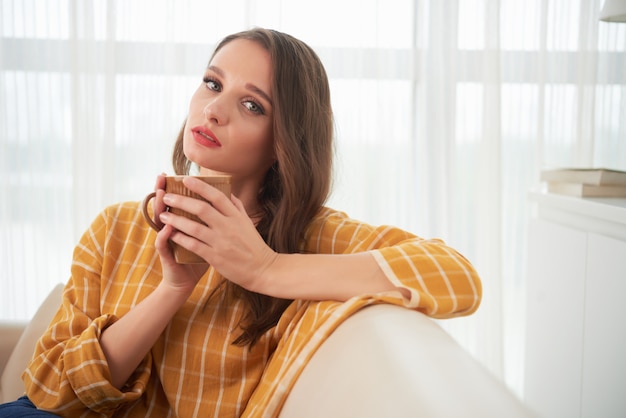 Schöne junge kaukasische Frau, die zu Hause auf Couch mit Tasse Tee aufwirft