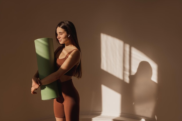 Schöne junge kaukasische Frau in Sportbekleidung, die Yogamatte hält, während sie drinnen steht Dunkelhaariges Mädchen bereitet sich auf das Training vor Gesunder Lebensstil Keep Fit Konzept