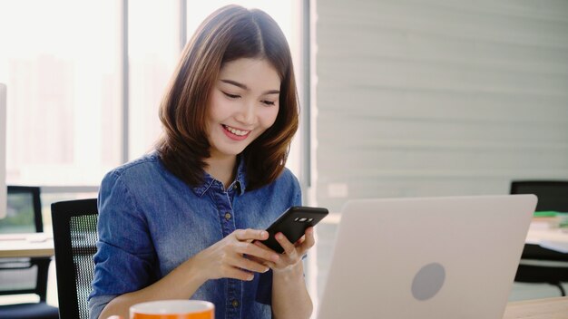 Schöne junge lächelnde asiatische Frau, die an Laptop beim Genießen der Anwendung des Smartphone im Büro arbeitet.