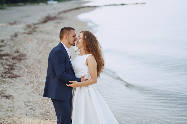 schöne junge langhaarige Braut im weißen Kleid mit ihrem jungen Ehemann auf dem Strand