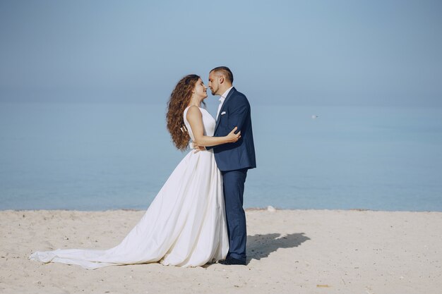 schöne junge langhaarige Braut im weißen Kleid mit ihrem jungen Ehemann auf dem Strand