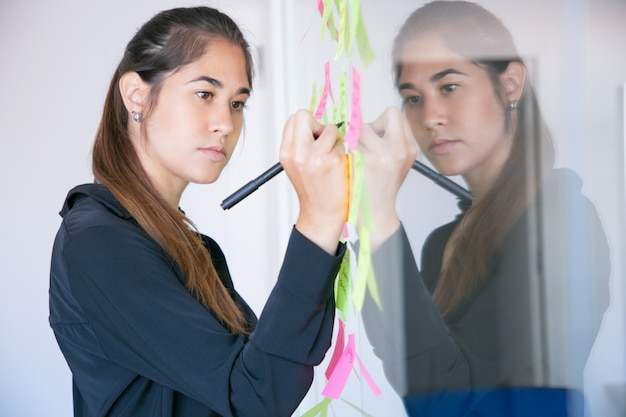 Schöne junge lateinische Geschäftsfrau, die auf Aufkleber mit Marker schreibt. Konzentrierte professionelle Managerin, die Ideen für das Projekt teilt und sich Notizen macht