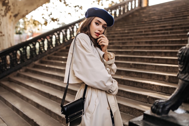 Schöne junge Pariserin mit brünetten Haaren in stylischer Baskenmütze, beigem Trenchcoat und schwarzer Tasche, die auf alten Treppen steht und sensibel im Freien posiert