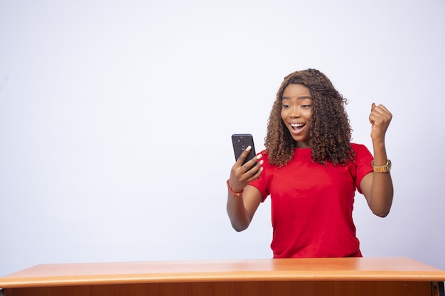 Schöne junge schwarze Frau sitzt an einem Schreibtisch, schaut auf ihr Telefon und fühlt sich aufgeregt