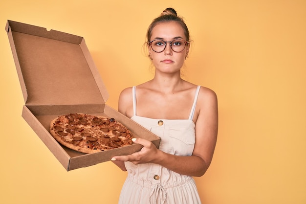 Schöne kaukasische Frau hält leckere Peperoni-Pizza in der Hand, entspannt mit ernstem Gesichtsausdruck, einfach und natürlich, während sie in die Kamera blickt