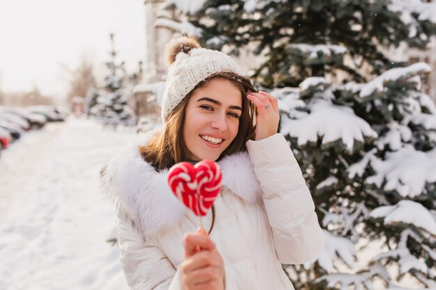 Schöne kaukasische Frau mit roter Süßigkeit, die Winter im Urlaub genießt. Außenfoto der entspannten Dame tragen weiße Strickmütze, die auf der Straße mit Schnee aufwirft