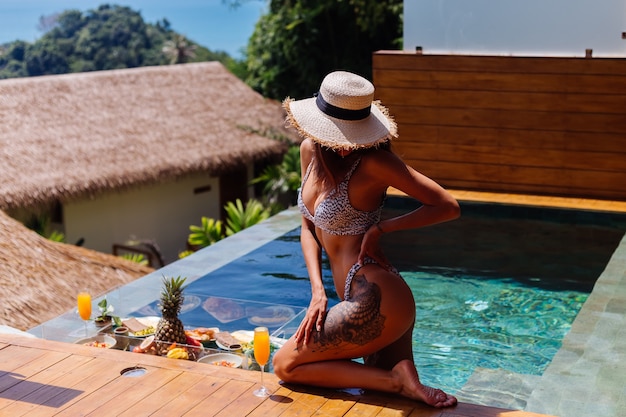 Schöne kaukasische gebräunte Frau im Bikini und im Strohhut mit schwimmendem Frühstück an der erstaunlichen Luxusvilla der Baliart am sonnigen Tag durch Schwimmbad, tropischer Hintergrund.