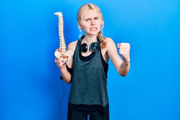 Kostenloses Foto schöne kaukasische sportlerin mit blonden haaren, die ein anatomisches modell der wirbelsäule hält, genervt und frustriert, schreit vor wut, schreit verrückt vor wut und erhobener hand