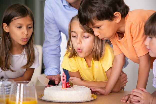 Schöne Kinder, die Kerze ausblasen und sich wünschen. Blondes kaukasisches Mädchen, das ihren Geburtstag mit Freunden und Kuchen feiert. Glückliche Kinder, die Spaß zusammen haben. Kindheits-, Feier- und Feiertagskonzept